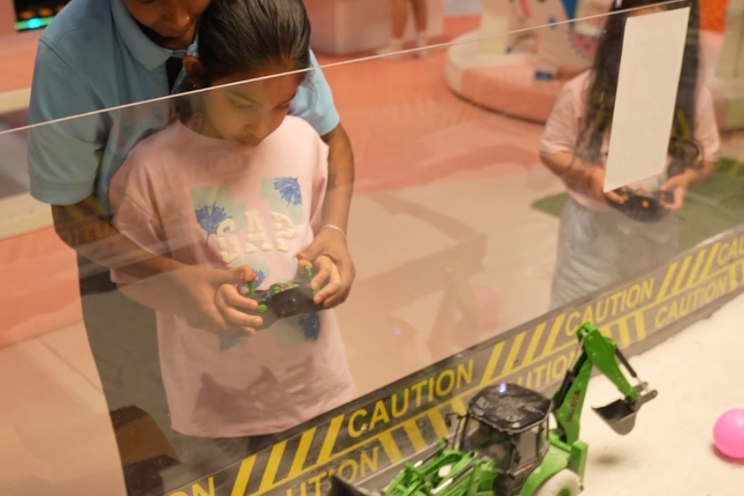 Children using remote controls to drive toy construction vehicles across a sand table